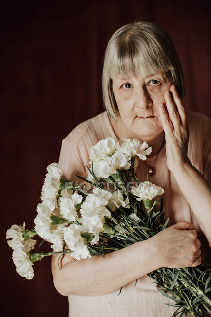 Dall'alto riflessivo vecchia femmina con i capelli grigi guardando la fotocamera e tenendo il bouquet di garofano bianco a casa — Foto stock