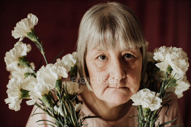 Von oben blickt eine nachdenkliche alte Frau mit grauen Haaren in die Kamera und hält einen Strauß weißer Nelken in der Hand. — Stockfoto
