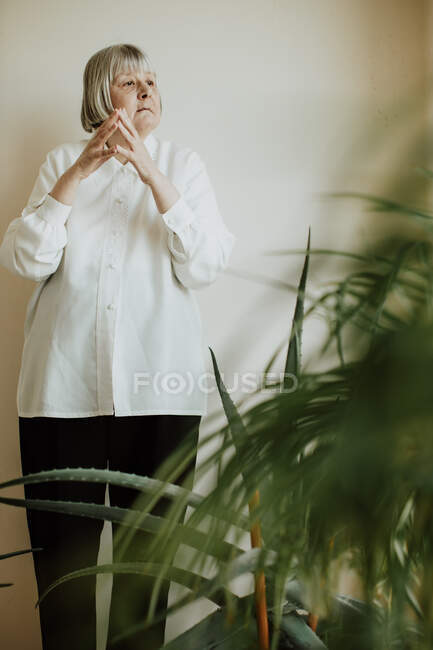 Сосредоточенная пенсионерка в белой блузке и черных брюках, стоящих у стены, делая жесты руками и отводя взгляд — стоковое фото