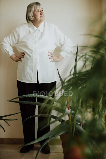 Женщина-пенсионер в белой блузке и черных брюках, стоящих у стены и смотрящих в сторону — стоковое фото