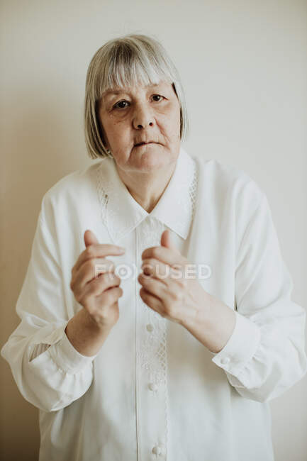 Traurige ältere Frau in weißer Bluse gestikuliert mit den Händen auf hellem Hintergrund und blickt in die Kamera — Stockfoto