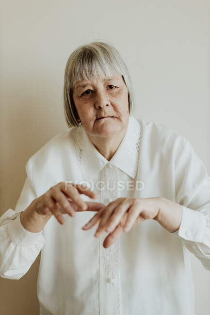 Sad elderly woman in white blouse gesturing with hands on light background looking at camera — Stock Photo