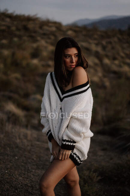 Romántica joven atractiva hembra con cabello oscuro usando suéter blanco y de pie en el campo en el crepúsculo - foto de stock