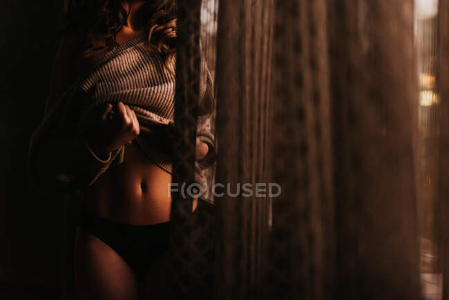 Cortar fêmea sedutora em lingerie preta e suéter mostrando barriga enquanto está de pé no escuro no quarto — Fotografia de Stock