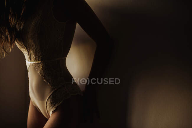 Crop slim female in white lace lingerie bodysuit leaning on wall in darkness — Stock Photo