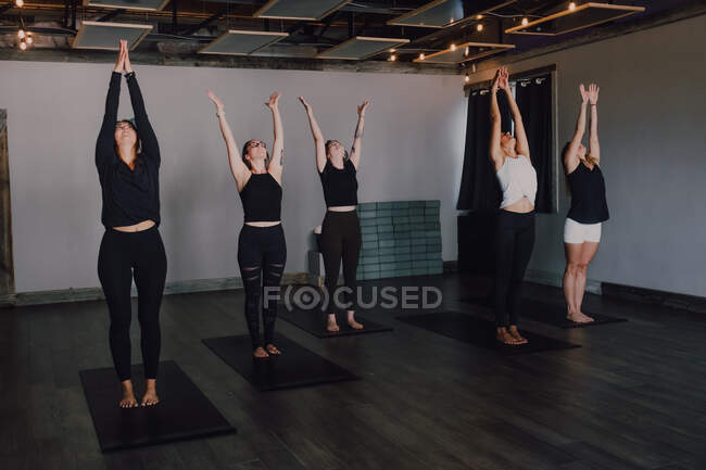 High angle of calm slender ladies in activewear with namaste standing in mountain position on sports mats on wooden floor and meditating in spacious contemporary gym looking up — Stock Photo