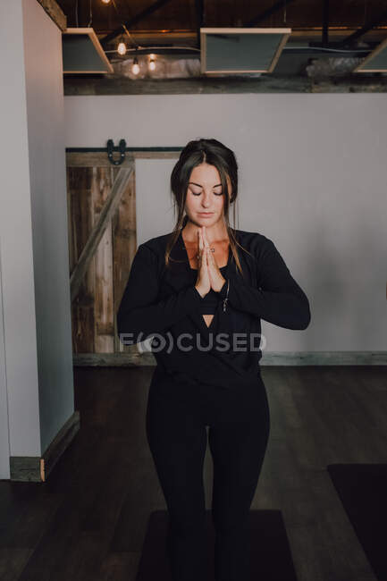 Ruhige, schlanke Frau in Aktivkleidung mit Namaste, die in Bergstellung auf Sportmatten auf Holzboden steht und mit geschlossenen Augen in einem geräumigen modernen Fitnessstudio meditiert — Stockfoto
