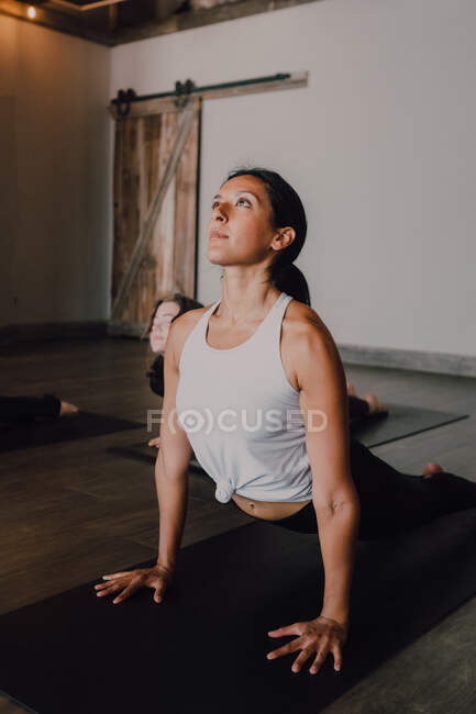 Barefoot unrecognizable woman in sportswear concentrating and doing upward facing dog exercise on sports mats on wooden floor against white walls of spacious hall — Stock Photo