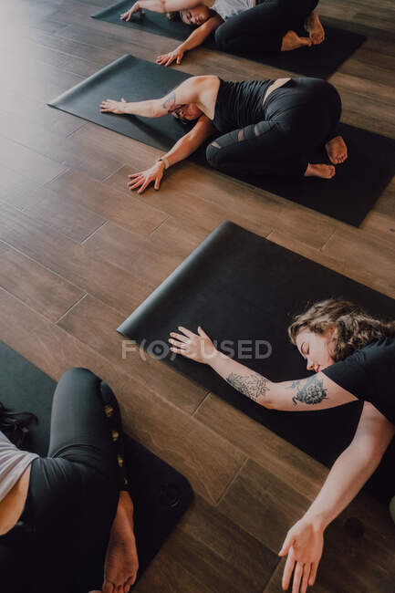 Сверху неузнаваемые дамы в спортивной одежде, сосредоточенные и лежащие в положении баласаны на спортивных ковриках на деревянном полу в просторной тренировочной комнате — стоковое фото