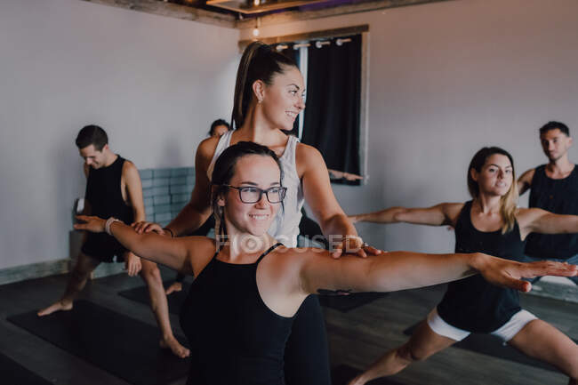 Smiling diligent female instructor in sportswear teaching warrior pose two group of sporty people doing exercise standing on sports mats in modern workout room — Stock Photo
