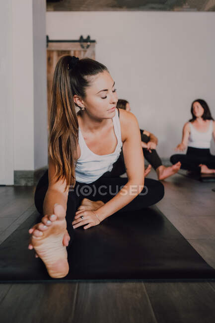 Diligent barefooted woman in sportswear performing head to knee forward bend exercise on mat while other sporty people resting after training in light contemporary gym — Stock Photo
