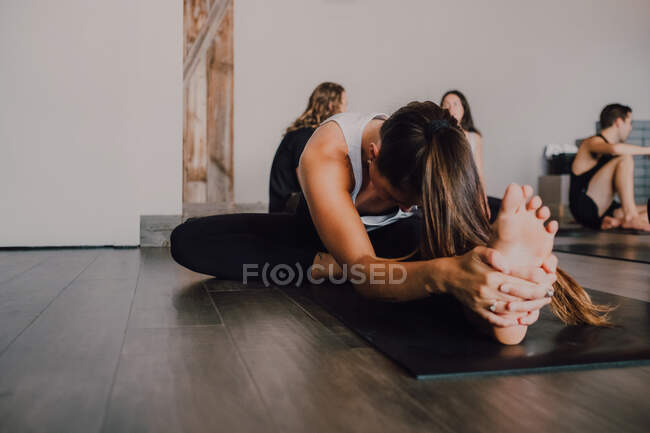 Diligent unrecognizable barefooted woman in sportswear performing head to knee forward bend exercise on mat while other sporty people resting after training in light contemporary gym — Stock Photo