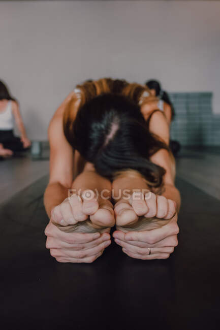 Mujer descalza irreconocible en ropa deportiva que estira el cuerpo mientras hace la pose de paschimottanasana sentada en la alfombra deportiva en el suelo en el gimnasio moderno - foto de stock