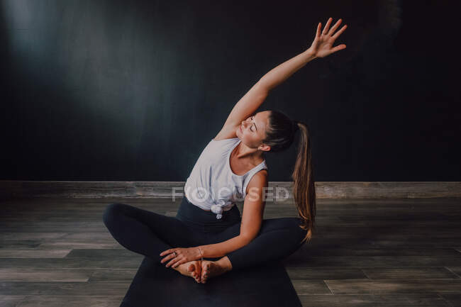 Young relaxed female with eyes closed and arm raised sitting in baddha konasana position on sports mat and practicing yoga in dark hall — Stock Photo