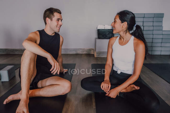 Young man and fit barefooted woman in sportswear smiling while looking at each other and talking while sitting on sports mats on floor and resting after yoga practice in contemporary studio — Stock Photo