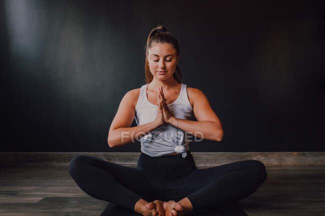 Calm relaxed female with eyes closed and namaste sitting in baddha konasana position on sports mat and concentrating while practicing yoga in dark modern studio — Stock Photo