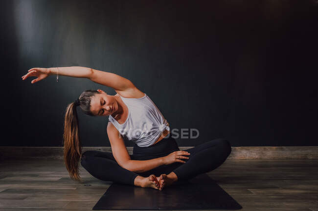 Jovem fêmea relaxada com os olhos fechados e braço levantado sentado na posição baddha konasana no tapete esportivo e praticando ioga no salão escuro — Fotografia de Stock
