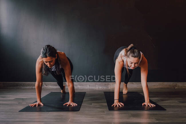 Босоногие женщины в спортивной одежде концентрируются и делают упражнения на доске на спортивных ковриках на деревянном полу против белых стен просторного зала — стоковое фото