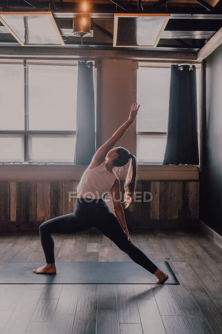 Vista posterior de la mujer irreconocible haciendo triángulo girado pose ejercicio de pie sobre alfombras deportivas en la sala de entrenamiento moderna - foto de stock