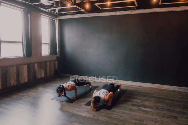 Зверху босоніж невпізнавані жінки в спортивному одязі концентрують і роблять дошку вправи на спортивних килимках на дерев'яній підлозі на білих стінах просторого залу — стокове фото