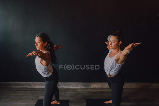 Mulheres em sportswear fazendo guerreiro posar dois exercício de ioga em pé em tapetes esportivos na sala de treino moderno — Fotografia de Stock