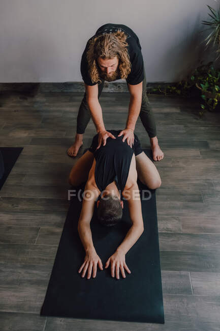 Жінка-інструктор у спортивному одязі навчає двох жінок, які лежать в позі баласани на спортивних килимках на дерев'яній підлозі у просторій тренувальній кімнаті — стокове фото