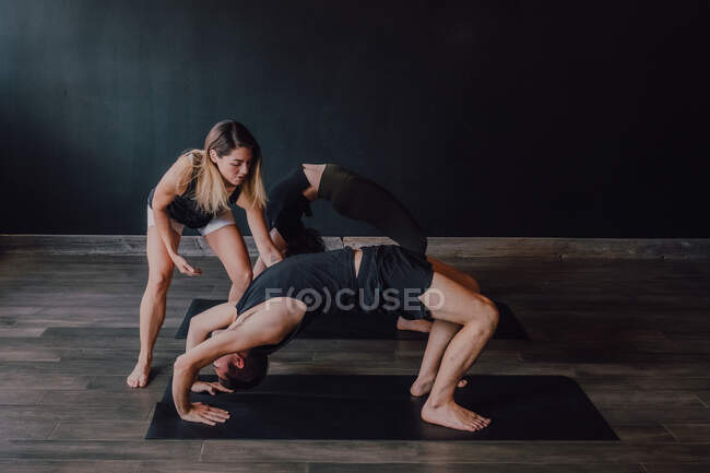 High angle of adult barefooted instructor in sportswear supporting man while teaching group of sporty people to do urdhva dhanurasana pose in contemporary workout room — Stock Photo
