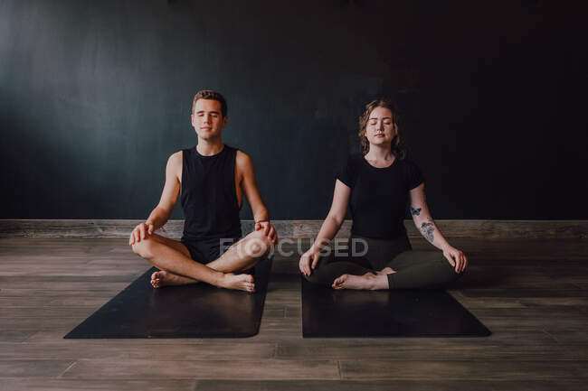 Jovem calmo e homem de sportswear com os olhos fechados e pernas cruzadas meditando juntos enquanto sentado na posição padmasana contra a parede traseira no estúdio de ioga contemporâneo — Fotografia de Stock