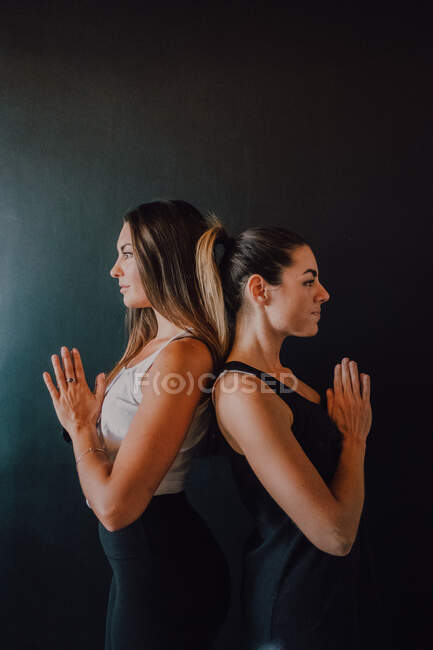 Side view of tranquil relaxed female athletes in activewear with hands folded practicing yoga in tadasana position while standing back to back and looking away against black wall in modern studio — Stock Photo