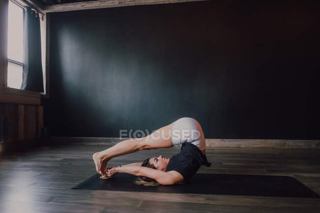 Vista lateral de ángulo alto de atleta femenina delgada enfocada en ropa deportiva que estira el cuerpo en posición halasana mientras entrena sola en un amplio estudio de yoga contemporáneo - foto de stock