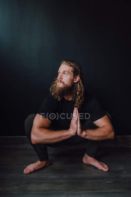Smiling handsome flexible male athlete in sportswear with namaste performing yoga kakasana position on wooden floor against black wall of contemporary workout room — Stock Photo