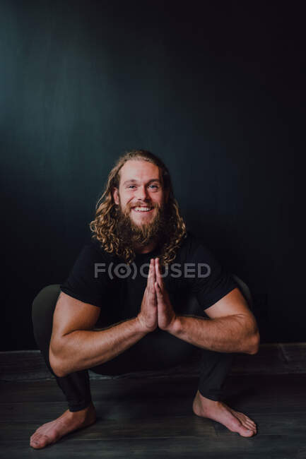 Sonriente guapo atleta masculino flexible en ropa deportiva con namaste realizando yoga kakasana posición en piso de madera contra pared negra de sala de entrenamiento contemporánea - foto de stock