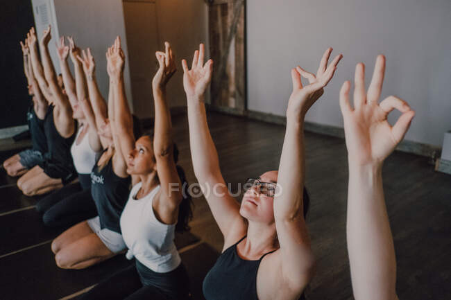 High angle of group of young sporty women and men in sportswear with arms raised and Gyan Mudra stretching body and meditating while sitting in virasana pose on sports mats and looking up training in modern yoga studio — Stock Photo