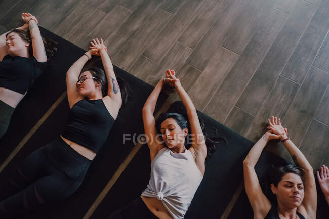Angle élevé de groupe de jeunes femmes et hommes sportifs en vêtements de sport avec les yeux fermés et les jambes croisées respiration relaxante et profonde tout en étant couché en position savasana et en méditant sur les tapis de sport dans le studio de yoga moderne — Photo de stock