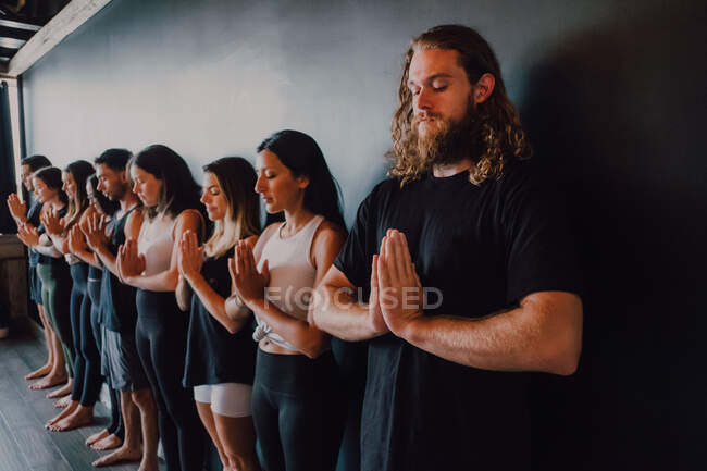 Gruppe fröhlicher junger Sportler in Aktivkleidung mit geschlossenen Augen, entspannt und tief atmend, während sie mit Namaste im Tadasana stehen und neben schwarzer Wand im modernen Yogastudio meditieren — Stockfoto