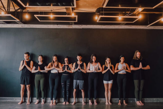 Group of happy young sporty people in activewear with eyes closed relaxing and deep breathing while standing with namaste in tadasana and meditating beside black wall in modern yoga studio — Stock Photo