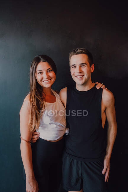 Attractive fit woman and happy millennial man in sportswear embracing and smiling at camera while standing against black wall — Stock Photo