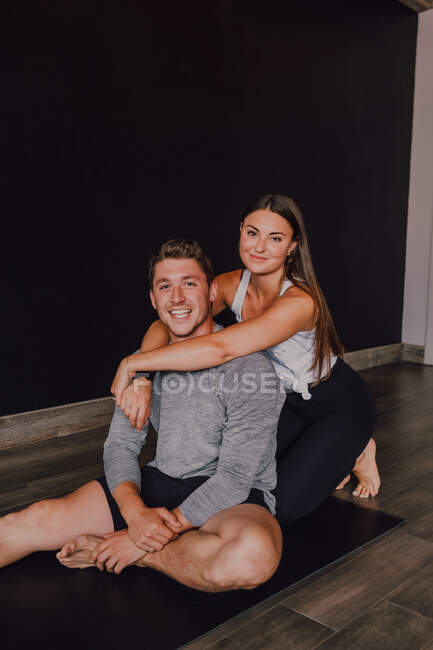 Hoher Winkel einer herrlich schlanken Frau und eines glücklichen erwachsenen Mannes in Sportbekleidung, der sich umarmt und in die Kamera lächelt, während er auf einer Sportmatte vor einer schwarzen Wand im modernen Yogastudio sitzt — Stockfoto