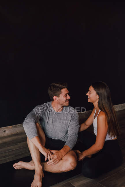 Herrliche schlanke Frau und glücklicher erwachsener Mann in Sportbekleidung umarmen und lächeln einander an, während sie auf der Sportmatte vor schwarzer Wand im modernen Yogastudio sitzen — Stockfoto
