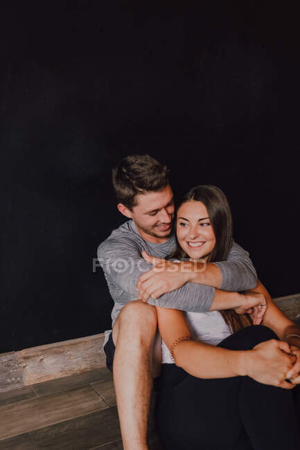 High angle of magnificent slim woman and happy adult man in sportswear embracing and smiling away while sitting on sports mat against black wall in contemporary yoga studio — Stock Photo