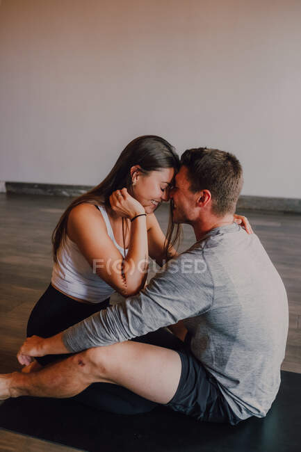 Hochwinkelseitenansicht einer glücklichen Frau und eines fröhlichen erwachsenen Mannes mit geschlossenen Augen in Activwear, die sich umarmen, während sie auf einer Sportmatte sitzen und sich in einem modernen Fitnessstudio amüsieren — Stockfoto