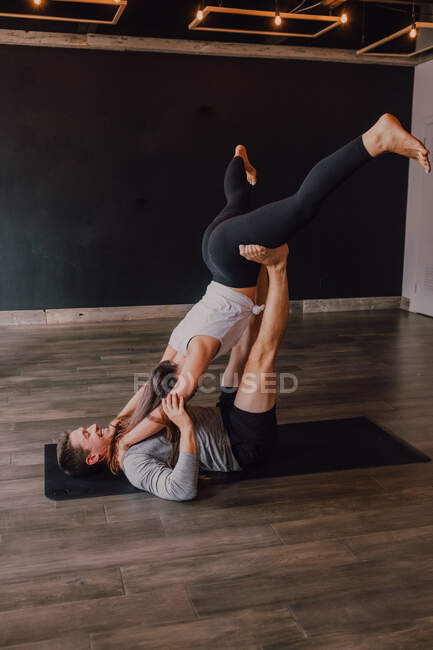 Vista laterale di uomo atletico gioioso con giovane partner femminile praticare acro yoga insieme in posa foglia piegata sul tappeto nero insieme in palestra moderna scura — Foto stock