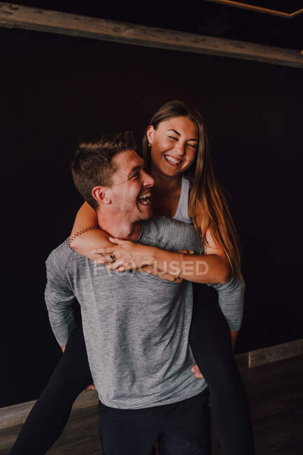 Excited adult man in sportswear piggybacking slender young woman while standing against black wall in contemporary gym — Stock Photo