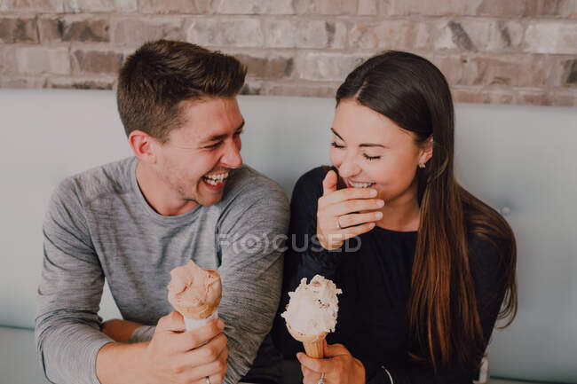 Hohe Winkel von gut gelaunten Mann und junge Frau in legerer Kleidung schauen einander mit leckerem Eis an, während sie am Tisch auf dem Sofa sitzen und es sich gemeinsam in einem modernen Café im Loft-Stil gemütlich machen — Stockfoto