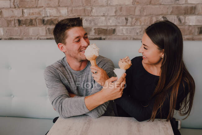 Високий кут веселого чоловіка і молодої жінки в повсякденному одязі дивлячись і годуючи один одного смачним морозивом, сидячи за столом на дивані і розслабляючись разом в сучасному кафе в стилі лофт — стокове фото