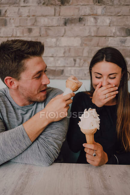 High angle of cheerful man and young woman in casual clothes looking at each other with tasty ice cream while sitting at table on sofa and relaxing together in modern cafe in loft style — Stock Photo