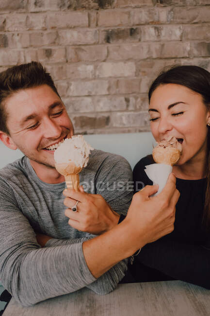 High angle of cheerful man and young woman in casual clothes looking and feeding each other with tasty ice cream while sitting at table on sofa and relaxing together in modern cafe in loft style — Stock Photo