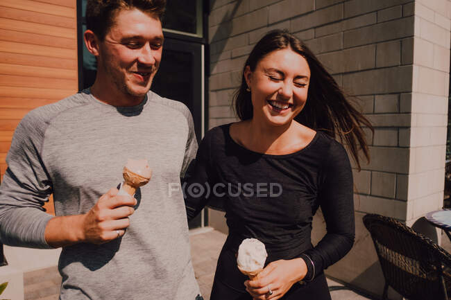 Crop adult man and positive fit woman with closed eyes in casual clothes having fun and laughing while walking along street and eating yummy ice cream — Stock Photo