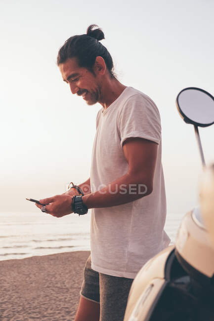 Seitenansicht positiver moderner, sportlicher Mann in T-Shirt und Shorts, der neben dem Fahrrad steht und bei Sonnenuntergang am Meer sein Handy benutzt — Stockfoto