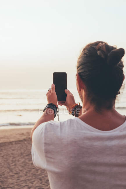 Vista posterior del hombre hipster en camiseta casual de pie en la playa y tomar una foto con el teléfono móvil mientras disfruta de la hermosa puesta de sol - foto de stock